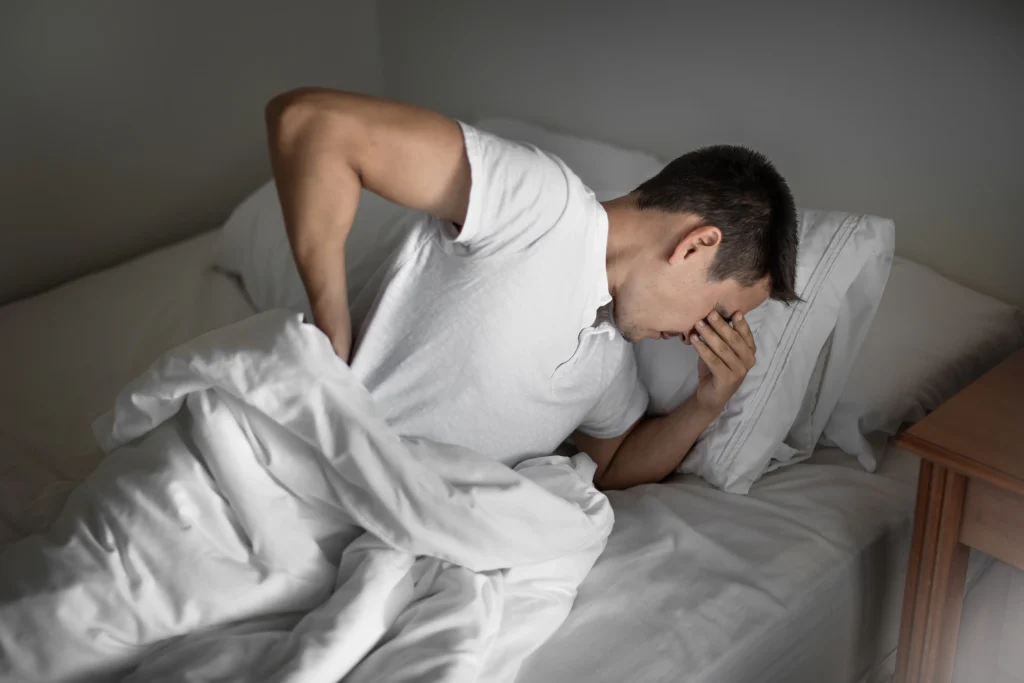 Homem com expressão de cansaço e dor, sentado na beira da cama com a mão no rosto e a outra na lombar, evidenciando o desconforto de não usar o travesseiro ideal para a coluna.
